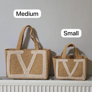 May include: Two tan crochet tote bags with white letter V detailing on the front. The larger bag is labeled Medium and the smaller bag is labeled Small. Both bags feature two top handles and a rectangular base, constructed from woven material.