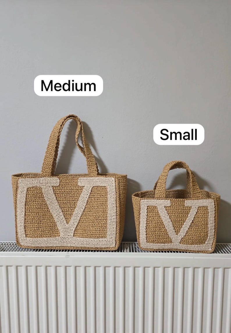 May include: Two tan crochet tote bags with white letter V detailing on the front. The larger bag is labeled Medium and the smaller bag is labeled Small. Both bags feature two top handles and a rectangular base, constructed from woven material.