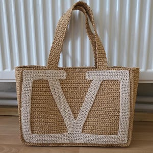 May include: A rectangular tote bag crafted from tan woven straw material. The front panel features a large, prominent letter V stitched in a contrasting cream-colored yarn. The bag includes two matching tan woven straw handles at the top.
