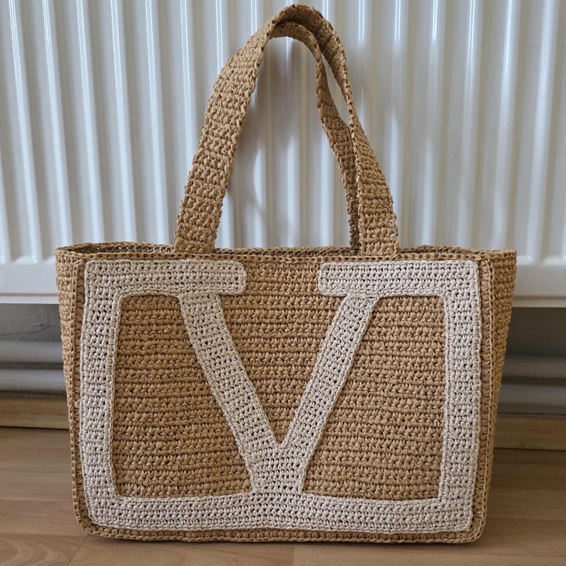May include: A rectangular tote bag crafted from tan woven straw material. The front panel features a large, prominent letter V stitched in a contrasting cream-colored yarn. The bag includes two matching tan woven straw handles at the top.