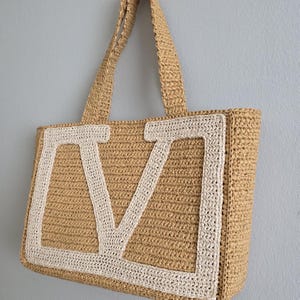 May include: A rectangular tote bag crafted from tan woven material featuring a large, cream-colored crocheted letter V centered on the front panel. The bag includes two matching tan woven shoulder straps and a structured, flat-bottomed design.