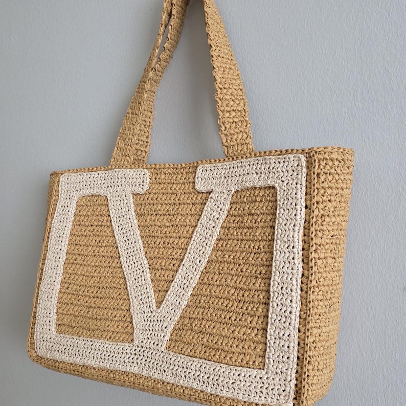 May include: A rectangular tote bag crafted from tan woven material featuring a large, cream-colored crocheted letter V centered on the front panel. The bag includes two matching tan woven shoulder straps and a structured, flat-bottomed design.