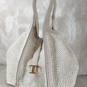 May include: Cream-colored woven tote bag with light beige leather accents. The bag features a structured design with a top handle and a decorative gold-tone buckle. The bag's woven texture and neutral color palette create a sophisticated look.