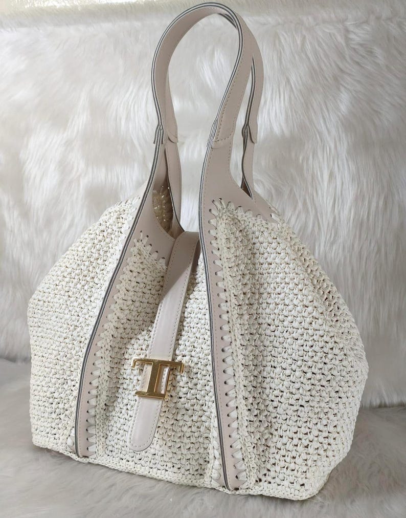May include: Cream-colored woven tote bag with light beige leather accents. The bag features a structured design with a top handle and a decorative gold-tone buckle. The bag's woven texture and neutral color palette create a sophisticated look.