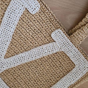 May include: A tan woven straw tote bag featuring a large, white, stitched letter V on the front panel. The bag is constructed from a textured, braided material with sturdy handles and a rectangular base, showcasing a clear contrast between the light tan body and the bright white letter applique.