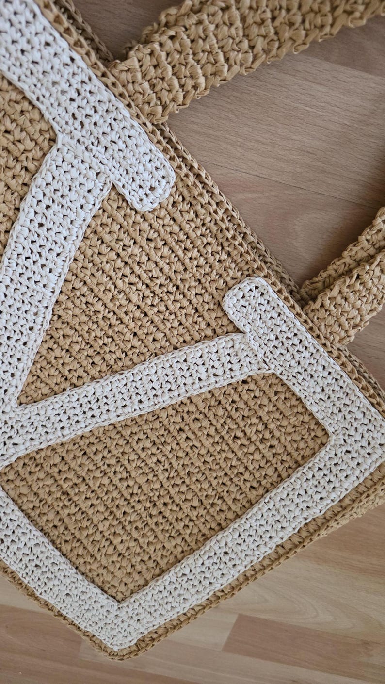 May include: A tan woven straw tote bag featuring a large, white, stitched letter V on the front panel. The bag is constructed from a textured, braided material with sturdy handles and a rectangular base, showcasing a clear contrast between the light tan body and the bright white letter applique.