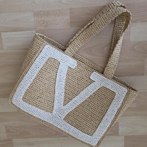 May include: A beige crocheted tote bag featuring a large, white, block-letter M stitched onto the front panel. The bag has two matching beige crocheted handles and a rectangular structure, crafted with a visible, textured, woven yarn pattern.