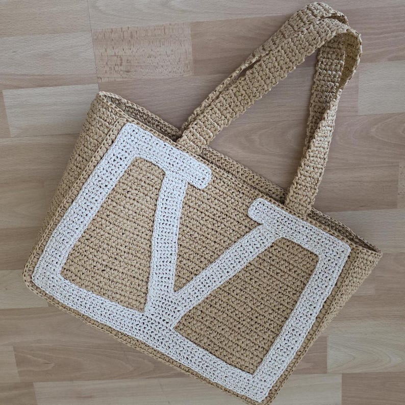 May include: A beige crocheted tote bag featuring a large, white, block-letter M stitched onto the front panel. The bag has two matching beige crocheted handles and a rectangular structure, crafted with a visible, textured, woven yarn pattern.