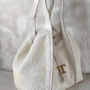 May include: Off-white woven tote bag with cream-colored leather handles and accents. The bag features a stitched design along the sides and a gold-colored buckle with the letter "T". The bag is designed with a spacious interior.