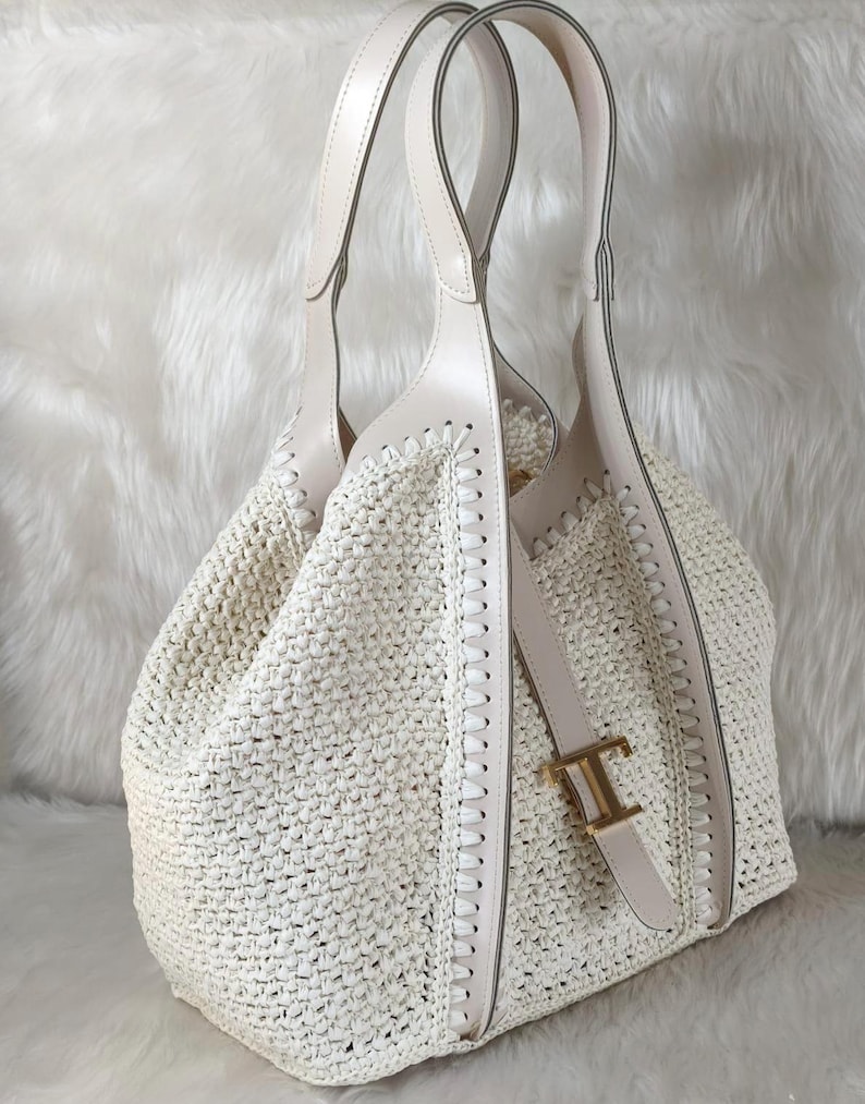 May include: Off-white woven tote bag with cream-colored leather handles and accents. The bag features a stitched design along the sides and a gold-colored buckle with the letter "T". The bag is designed with a spacious interior.