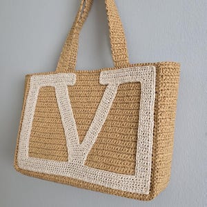May include: A rectangular tote bag crafted from tan woven material, featuring two matching shoulder straps. The front of the bag displays a large, prominent letter V appliqu&eacute;d in a contrasting cream-colored woven material with a thick border.