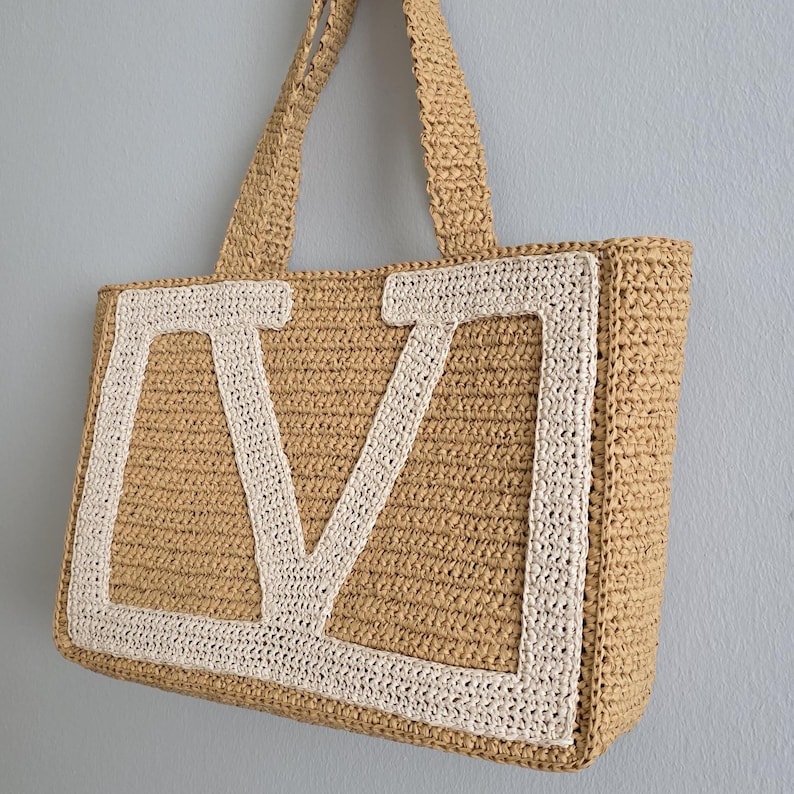 May include: A rectangular tote bag crafted from tan woven material, featuring two matching shoulder straps. The front of the bag displays a large, prominent letter V appliqu&eacute;d in a contrasting cream-colored woven material with a thick border.