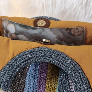 May include: A handmade handbag with a crocheted front in a rainbow of colors, including brown, blue, purple, and yellow. The bag has a mustard-colored interior and a silver crocheted handle. The interior features a floral print pocket and a magnetic clasp.