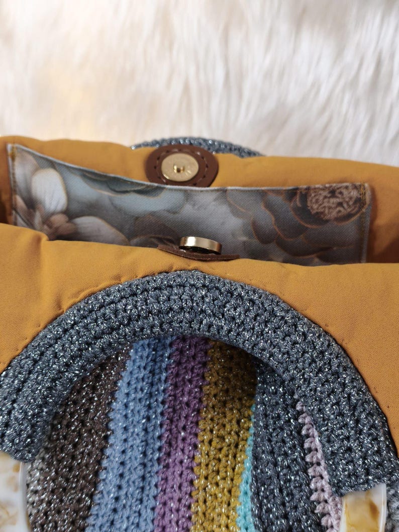 May include: A handmade handbag with a crocheted front in a rainbow of colors, including brown, blue, purple, and yellow. The bag has a mustard-colored interior and a silver crocheted handle. The interior features a floral print pocket and a magnetic clasp.