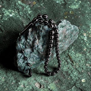 Peut inclure: Un chapelet composé de perles sombres sphériques ornées de rayures blanches verticales. Les perles sont reliées par un fin cordon noir, se terminant par une pièce de pompon noire lisse et allongée. Le chapelet est drapé sur une pierre texturée.