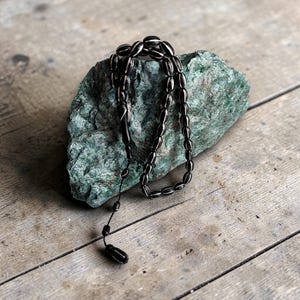 Peut inclure: Un chapelet noir avec des perles ovales et des accents argentés repose sur une pierre verte. Les perles sont reliées par un cordon noir. La pierre est posée sur une surface en bois vieilli.