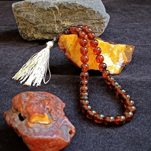May include: A string of translucent, deep amber-colored prayer beads featuring a silver-toned metal accent and a long, white tassel. The beads are arranged in a loop, resting against a large, jagged piece of raw, golden-yellow amber.