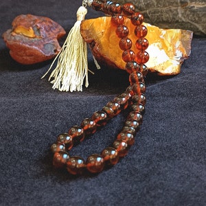 May include: A string of polished, translucent dark amber beads arranged in a loop, featuring a silver-toned metal connector and a light cream-colored fabric tassel. The beads exhibit a deep reddish-brown hue with natural internal variations.