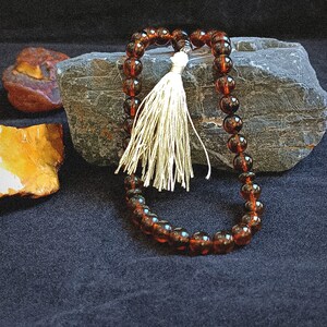 May include: A string of translucent, dark amber-colored spherical beads arranged in a loop, featuring a single off-white decorative tassel attached to the top. The beads are smooth and polished, reflecting light across their rounded surfaces.