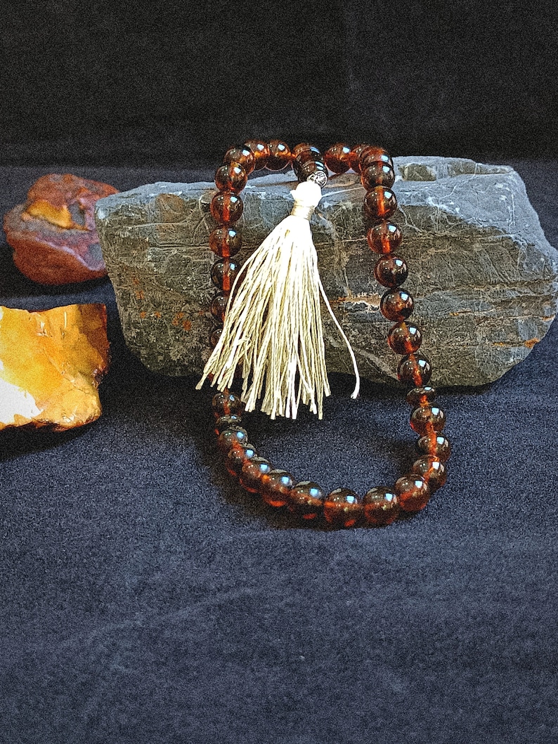 May include: A string of translucent, dark amber-colored spherical beads arranged in a loop, featuring a single off-white decorative tassel attached to the top. The beads are smooth and polished, reflecting light across their rounded surfaces.