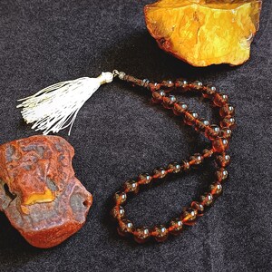 May include: A string of dark amber prayer beads featuring smooth, translucent, round beads connected by a cord. The prayer beads are finished with a decorative white tassel at one end, positioned near two pieces of raw, unpolished amber stone.