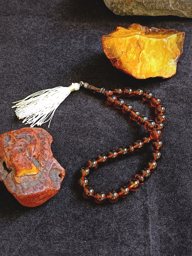 May include: A string of dark amber prayer beads featuring smooth, translucent, round beads connected by a cord. The prayer beads are finished with a decorative white tassel at one end, positioned near two pieces of raw, unpolished amber stone.