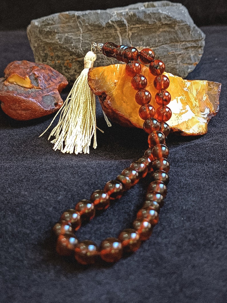 May include: A string of translucent, deep amber-colored prayer beads featuring a polished, spherical design. The strand is finished with a decorative, light-colored tassel attached by a small silver-toned metal component at the top of the loop.
