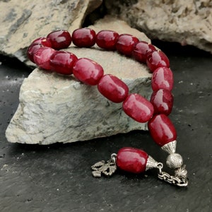 May include: A red gemstone prayer beads necklace with silver accents. The beads are oval shaped and have a smooth, polished finish. The necklace has a silver tassel and a silver clasp.