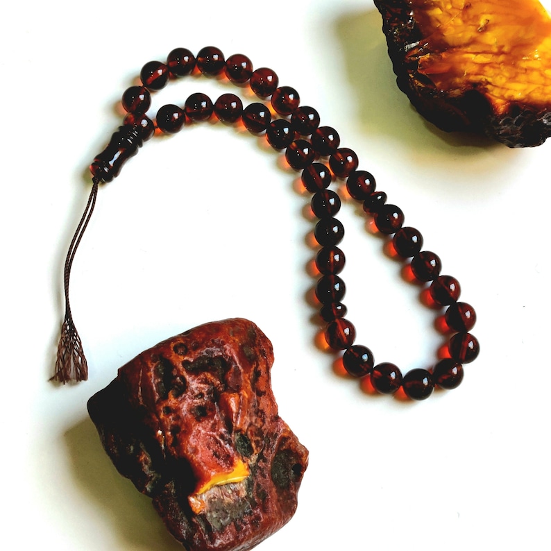 May include: A string of dark reddish-brown translucent amber prayer beads arranged in a loop, featuring a matching carved bead and a brown tassel at one end, positioned near two pieces of raw, unpolished amber with deep orange and brown tones.