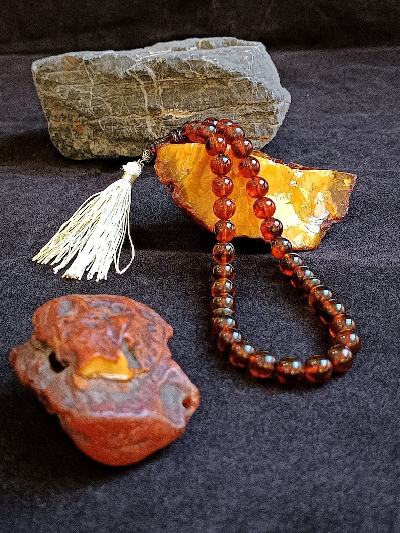 May include: A string of translucent, deep amber-colored prayer beads featuring a silver-toned metal accent and a long, white tassel. The beads are arranged in a loop, resting against a large, jagged piece of raw, golden-yellow amber.