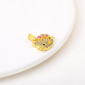 May include: A gold-colored pendant in the shape of a cartoon character's head. The pendant features a pink bow, yellow accents, and is encrusted with clear crystals. The pendant is on a white surface.