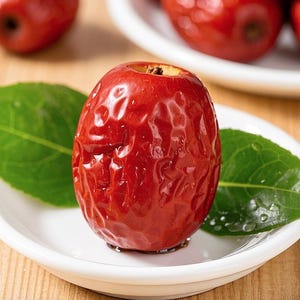 Op de afbeelding: Een enkele rode jujube, ook wel Chinese dadel genoemd, op een wit bord met groene bladeren. De jujube heeft een gerimpelde textuur en een glanzende afwerking.