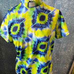 May include: A short-sleeved shirt with a high collar and a vibrant tie-dye pattern. The shirt features a repeating design of blue and yellow circular patterns. The collar has a white insert.