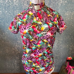 May include: A short-sleeved shirt with a vibrant, colorful animal print design. The shirt features a high collar and is displayed on a mannequin. The print includes shades of pink, purple, green, and yellow, with black accents.