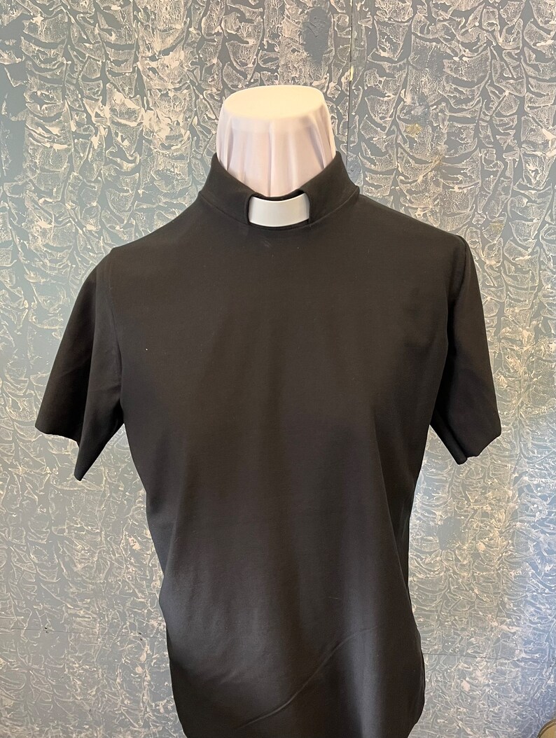 May include: A black short-sleeved clergy shirt featuring a high mock neck collar with a white tab insert. The shirt is displayed on a mannequin torso, showcasing the solid black fabric and the distinct white clerical collar detail at the neckline.