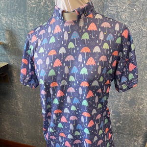 May include: A short-sleeved shirt with a navy blue background and a pattern of colorful umbrellas. The umbrellas are in shades of pink, orange, green, and blue. The shirt has a high collar.