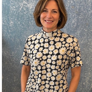 May include: A short-sleeved shirt featuring a repeating pattern of white daisies with yellow centers on a black background. The shirt has a high mock neck collar with a white rectangular tab insert at the center front, creating a clerical style.