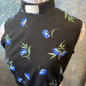 May include: Black top with a high collar and a floral pattern of blue flowers and green leaves. The top is worn over a white garment, creating a layered effect. The floral design adds a touch of color and detail to the garment.