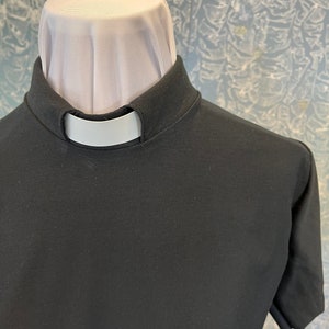May include: A black short-sleeved clergy shirt featuring a high stand-up collar with a white tab insert. The shirt is constructed from a solid black fabric and includes a white fabric neck piece visible above the collar line.