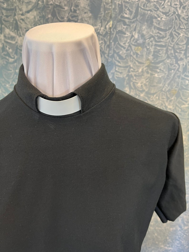 May include: A black short-sleeved clergy shirt featuring a high stand-up collar with a white tab insert. The shirt is constructed from a solid black fabric and includes a white fabric neck piece visible above the collar line.