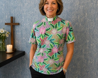 May include: A short-sleeved clergy shirt featuring a vibrant, multi-colored botanical print with green leaves and pink floral patterns on a geometric background, finished with a white clerical collar insert at the high neckline.
