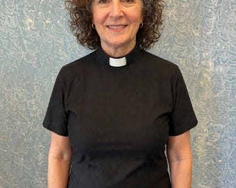 May include: A black short-sleeved shirt featuring a high mock neck collar with a white rectangular clerical tab insert at the center front. The garment is constructed from a solid black fabric and is designed with a standard fit, short sleeves, and a clean, finished neckline that accommodates the removable white clerical tab.