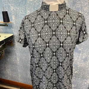 May include: A short-sleeved shirt with a high collar and a white tab. The shirt features a black and white ornate pattern. The shirt is displayed on a mannequin.