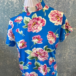 May include: A short-sleeved, blue shirt with a high collar, adorned with a vibrant floral pattern. The design features large pink and white flowers with green leaves, creating a bold and colorful aesthetic.