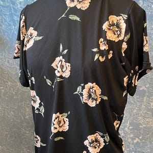 May include: Black short-sleeved top with a mock neck and a floral pattern. The flowers are peach and pink with green leaves. The top is displayed on a mannequin.