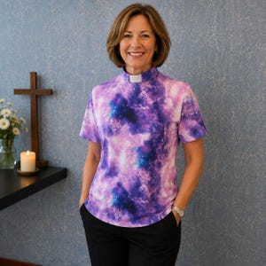 May include: A short-sleeved clergy shirt featuring a vibrant purple, blue, and pink galaxy print pattern. The shirt has a high mock neck collar with a white clerical tab insert at the center front, providing a distinct contrast against the cosmic design.