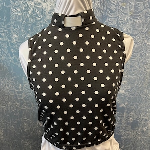 May include: A black sleeveless top with white polka dots. The top has a high, rounded collar with a small opening at the back. The bottom hem is elasticized. The top is displayed on a mannequin.