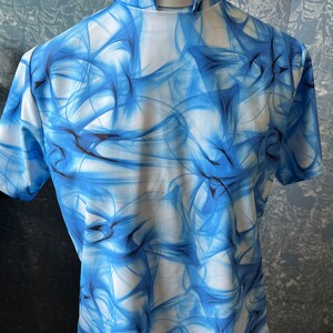 May include: A short-sleeved shirt with a high collar, featuring a blue and white abstract design. The pattern resembles flowing smoke or water, creating a dynamic visual effect. The shirt is displayed on a mannequin.