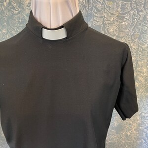 May include: A black short-sleeved clergy shirt featuring a high stand-up collar with a white tab insert. The garment is displayed on a mannequin, showcasing the solid black fabric and the distinct white rectangular clerical collar insert at the neck.