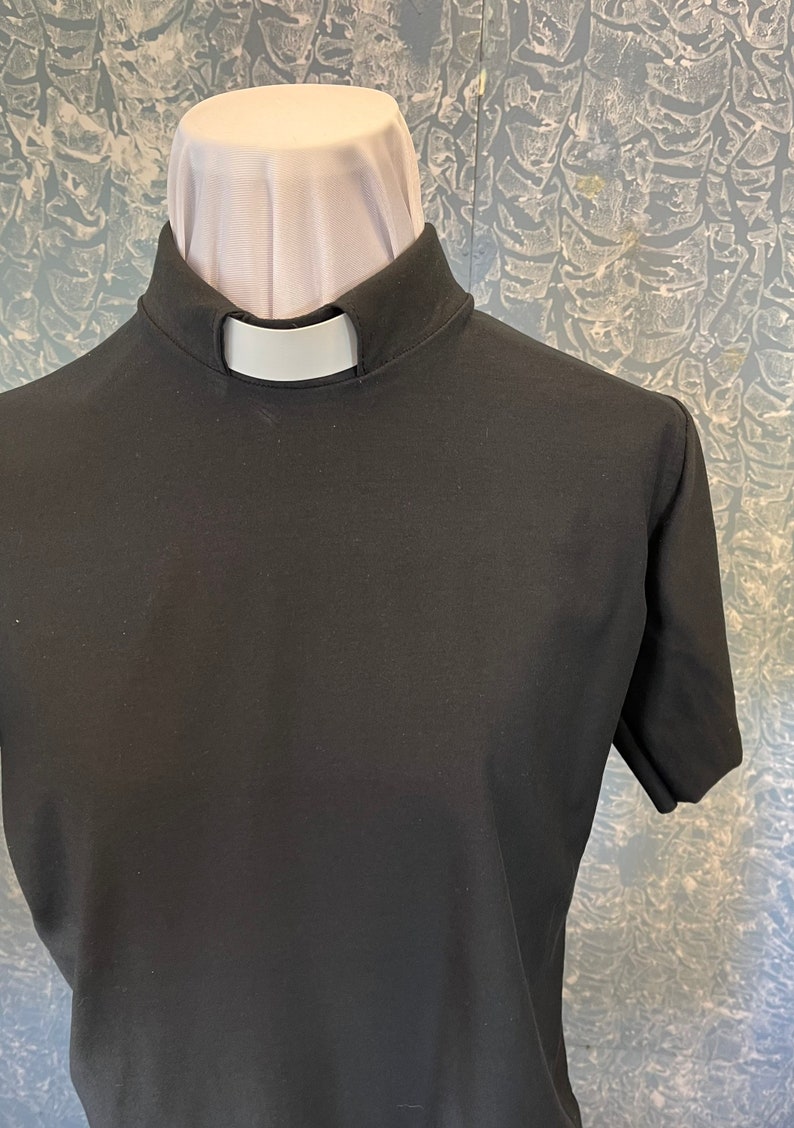 May include: A black short-sleeved clergy shirt featuring a high stand-up collar with a white tab insert. The garment is displayed on a mannequin, showcasing the solid black fabric and the distinct white rectangular clerical collar insert at the neck.